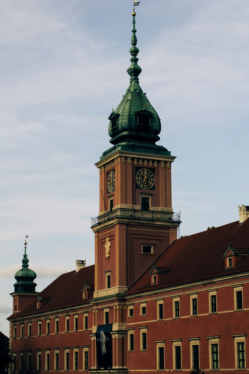 The terracotta facade and green-domed clock tower of the Royal Castle in Warsaw against a pale evening sky.
